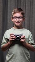 vertical boy in glasses holds old vintage camera in hands. looking into camera, medium shot. concept beginner photographer. child learns to take pictures. portrait of boy with old vintage camera video