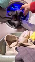 vertical POV person loads clothes into a washing machine in well-organized laundry room at home, with shelves and detergent bottles. Blue light illuminates the scene. Busy housekeeper loading clothes video