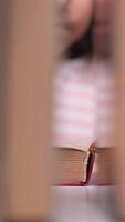 vertical close-up little girl leafing through a book in the library. Stacks of books on the table surround the child. The little girl reads stories from the book with interest video