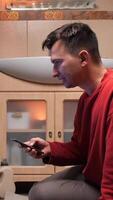 vertical Man is kneeling in the bathroom, using a smartphone while waiting for the washing machine to finish its cycle, with towels hanging on the side. texting video