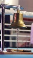 vertical Brass boxing bell hanging from a metal stand with a mallet resting on the wooden base, placed on a blue canvas-covered boxing ring corner video