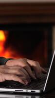 vertical Man is sitting by the fireplace and typing on a laptop. He is wearing a sweater and his hands are illuminated by the light of the screen video