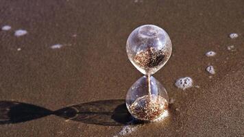 Glass sand timer stands on beach with transparent sea waves video