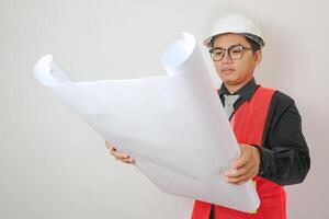 Portrait young asian man is engineer wearing helmet holding and looking blueprint with confident isolated on white background, handsome male is architect or contractor, worker or labor concept. photo