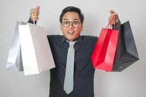 A portrait of an Indonesian Asian man wearing a gray outfit, looking happy and posing while holding shopping bags, isolated on a white background. photo