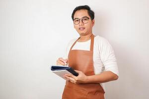 Smiling young Asian man barista barman employee wearing brown apron working in coffee shop write down order menu in notebook isolated on white background. Small business startup concept photo