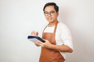 Smiling young Asian man barista barman employee wearing brown apron working in coffee shop write down order menu in notebook isolated on white background. Small business startup concept photo