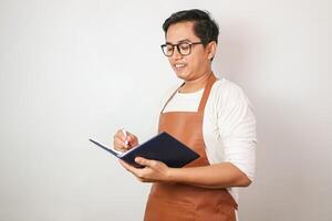Asian barista man in blue apron taking order, smiling and writing on menu book list isolated image on white background photo