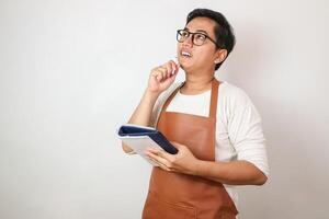 Asian barista man in brown apron taking order, writing on menu book list, thinking with pen on head. Isolated image on white background photo