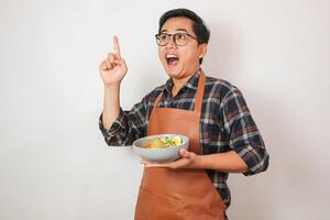 Portrait asian young man wearing an apron and holding a bowl of food is posing against a white background. photo