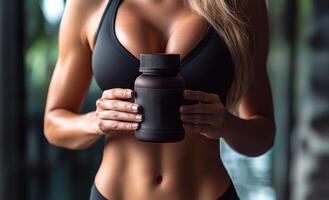 A woman holding a bottle of pills in her hand. The bottle is black and has a label on it. The woman is wearing a black tank top and has a toned body. Concept of health and fitness photo