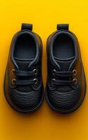 Two small black shoes with laces are placed on a yellow surface. The shoes are shiny and appear to be made of leather. The yellow background contrasts with the black shoes, making them stand out photo