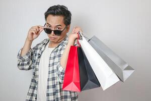Young asian man surprised and surprised, lowering glasses looking while holding shopping bags while looking at camera with open mouth isolated on plain white background studio portrait. photo