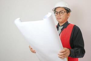 Asian engineer handsome man or architect looking forward holding paperwork blueprint with white safety helmet in construction site. Standing at modern building. photo