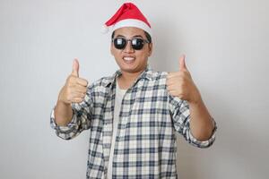 Cheerful young Asian man in Santa hat showing thumbs up, looking at camera on white studio background. Happy New Year celebration merry holiday concept. photo