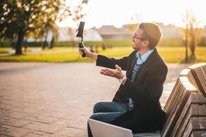 Businessman vlogger in coat wearing eyeglasses and using mobile phone with gimbal. He is talking while recording vlog in the park on a sunny autumn afternoon. photo