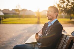 Portrait of content young businessman with arms crossed looking away while sitting on the bench in public park at the sunset. He is resting and thinking about something. photo