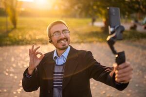 Businessman vlogger in coat wearing in-ear headphones and eyeglasses showing ok sign while using mobile phone with gimbal. He is standing in the park and recording vlog on a sunny autumn afternoon. photo