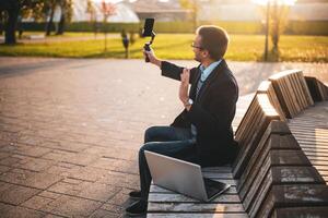 Businessman vlogger in coat wearing eyeglasses and waving while using mobile phone with gimbal. He is sitting on a bench in the park with laptop and recording vlog on a sunny autumn afternoon. photo