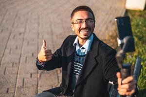 Cheerful businessman vlogger wearing eyeglasses and showing thumb up while using mobile phone with gimbal. He is sitting on a bench and recording vlog outdoor. photo