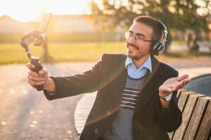 Businessman vlogger in coat wearing headphones and eyeglasses. He is using mobile phone with gimbal in the park and talking while recording vlog on a sunny autumn afternoon. photo