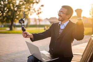 Excited businessman vlogger in coat wearing eyeglasses and gesturing while using mobile phone with gimbal for recording vlog. He is sitting on a bench in the park on a sunny autumn afternoon. photo