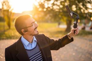 Smiling businessman vlogger in coat wearing in-ear headphones and eyeglasses using mobile phone with gimbal for recording vlog while standing in the park on a sunny autumn afternoon. photo