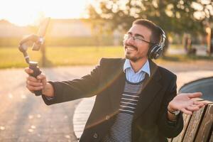 Businessman vlogger in coat wearing headphones and eyeglasses. He is using mobile phone with gimbal in the park and talking while recording vlog on a sunny autumn afternoon. photo