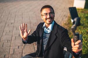 Cheerful businessman vlogger wearing eyeglasses and waving while using mobile phone with gimbal. He is sitting on a bench and recording vlog outdoor. photo