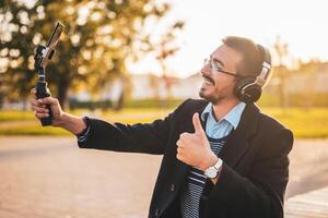 Businessman vlogger in coat wearing headphones and eyeglasses. He is showing thumb up and using mobile phone with gimbal while recording vlog on a sunny autumn afternoon in the park. photo