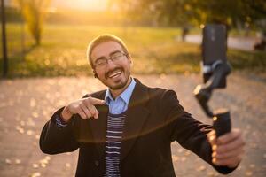 Cheerful businessman vlogger in coat wearing in-ear headphones and eyeglasses while pointing at mobile phone with gimbal. He is standing in the park and recording vlog on a sunny autumn afternoon. photo