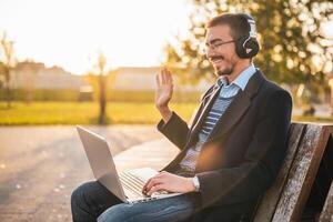 Happy businessman in coat with eyeglasses and headphones waving while using laptop and sitting in the park on a sunny autumn afternoon. photo