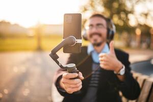 Businessman vlogger is using mobile phone with gimbal and showing thumb up while recording vlog outdoor. He is wearing headphones and eyeglasses. Focus on the phone and gimbal. photo