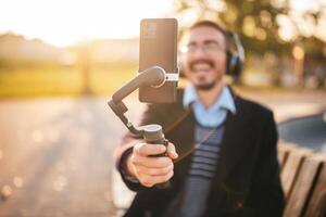 Businessman vlogger is using mobile phone with gimbal while sitting outdoor and recording vlog on a sunny autumn afternoon. He is wearing headphones and eyeglasses. Focus on the phone and gimbal. photo