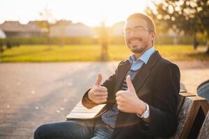 Portrait of successful businessman with eyeglasses showing thumbs up while sitting on a bench in public park at sunset. photo