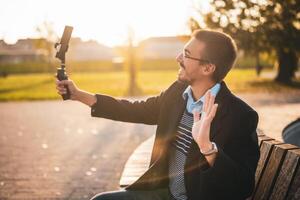 Happy businessman vlogger in coat wearing eyeglasses and using mobile phone with gimbal. He is sitting on a bench in the park and waving while recording vlog on a sunny autumn afternoon. photo