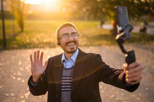 Happy businessman vlogger in coat wearing in-ear headphones and eyeglasses waving while using mobile phone with gimbal. He is standing in the park and recording vlog on a sunny autumn afternoon. photo