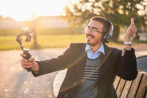 Businessman vlogger in coat wearing headphones and eyeglasses. He is using mobile phone with gimbal in the park and talking while recording vlog on a sunny autumn afternoon. photo