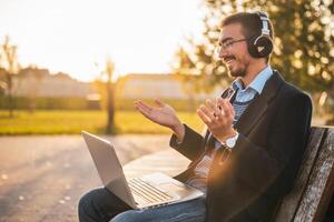 Smiling businessman in coat with eyeglasses and headphones using laptop and talking while sitting in the park on a sunny autumn afternoon. photo