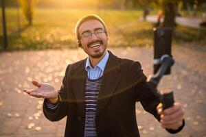 Young businessman vlogger in coat wearing in-ear headphones and eyeglasses talking while using mobile phone with gimbal. He is standing in the park and recording vlog on a sunny autumn afternoon. photo