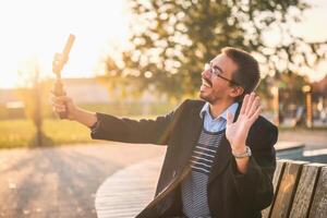 Happy businessman vlogger in coat wearing eyeglasses and using mobile phone with gimbal. He is sitting on a bench in the park and waving while recording vlog on a sunny autumn afternoon. photo