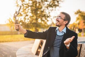 Businessman vlogger in coat wearing eyeglasses and using mobile phone with gimbal. He is sitting on a bench in the park and talking while recording vlog on a sunny autumn afternoon. photo