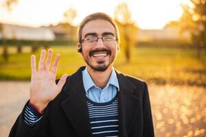 Close up portrait of happy businessman in coat with eyeglasses using in-ear headphones and waving while looking at camera. He is standing in the park on a sunny autumn afternoon. photo