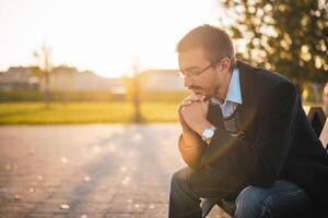 Sad businessman with eyeglasses sitting on the bench in public park at the sunset and thinking. photo