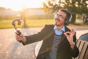 Businessman vlogger in coat wearing headphones and eyeglasses. He is showing ok sign and using mobile phone with gimbal while recording vlog on a sunny autumn afternoon in the park. photo