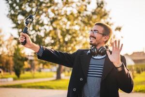 Businessman vlogger in coat wearing eyeglasses and headphones. He is using mobile phone with gimbal in the park and waving while recording vlog on a sunny autumn afternoon. photo