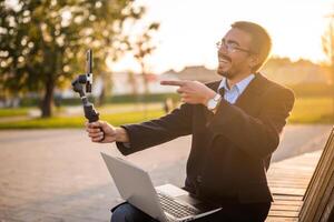 Cheerful businessman vlogger in coat wearing eyeglasses and pointing at mobile phone with gimbal. He is sitting on a bench in the park and recording vlog on a sunny autumn afternoon. photo
