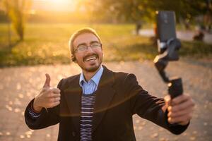Businessman vlogger in coat wearing in-ear headphones and eyeglasses showing thumb up while using mobile phone with gimbal. He is standing in the park and recording vlog on a sunny autumn afternoon. photo