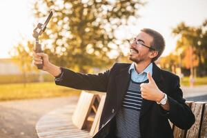 Businessman vlogger in coat wearing eyeglasses and showing thumb up while using mobile phone with gimbal. He is sitting on a bench in the park and recording vlog on a sunny autumn afternoon. photo