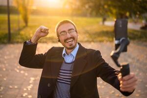 Excited businessman vlogger in coat wearing in-ear headphones and eyeglasses gesturing while using mobile phone with gimbal. He is standing in the park and recording vlog on a sunny autumn afternoon. photo